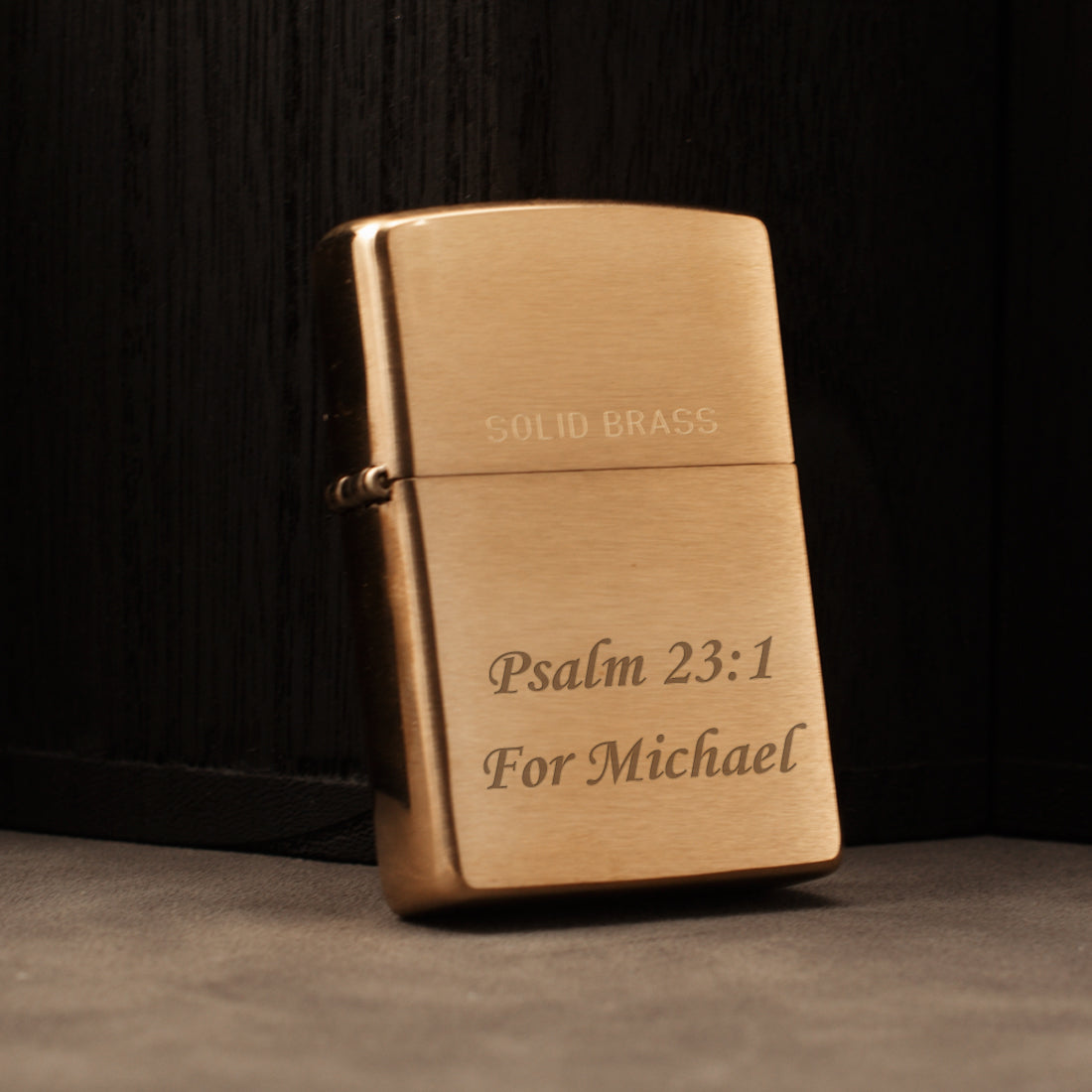 A solid brass lighter with engraved text reading 'PSALM 23:1 For Michael' on a dark wood background.