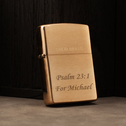 A solid brass lighter with engraved text reading 'PSALM 23:1 For Michael' on a dark wood background.