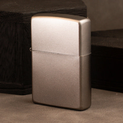A classic rectangular lighter with a brushed metallic finish, standing upright on a textured surface with dark wooden blocks in the background.