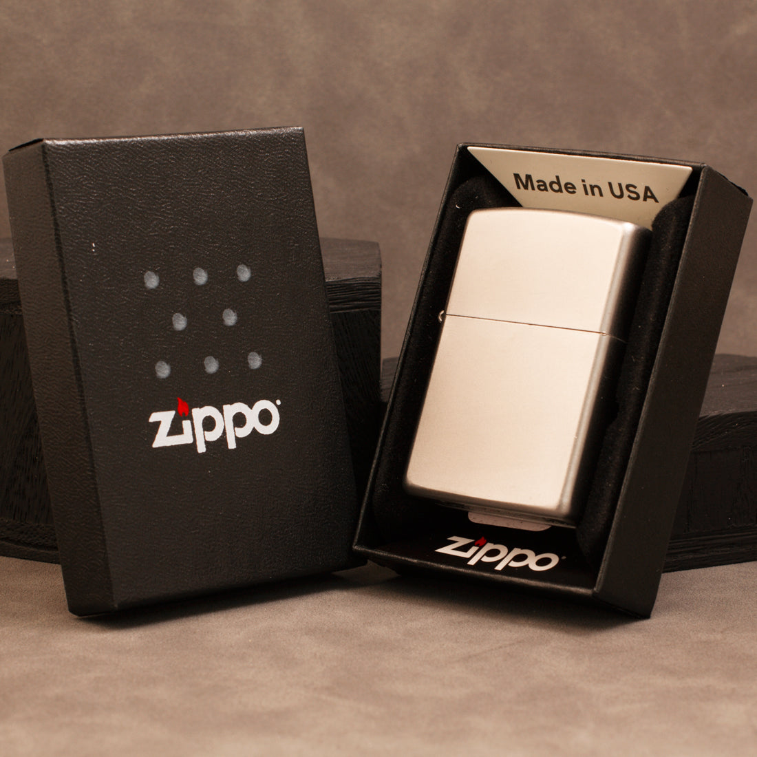A silver Zippo lighter displayed in an open black Zippo box next to its closed black presentation box on a textured brown background.