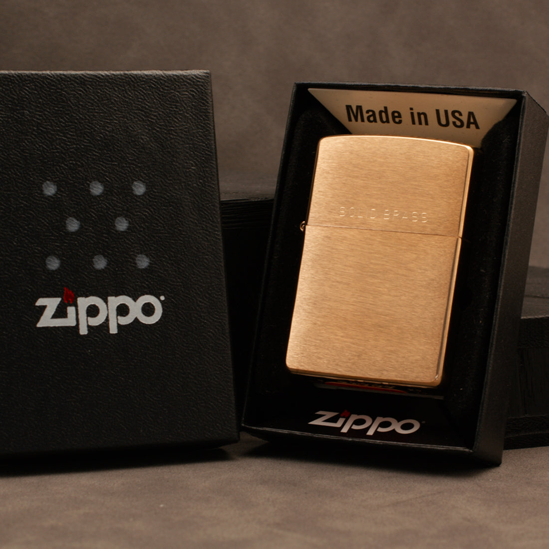 A brushed gold Zippo lighter displayed inside an open black Zippo presentation box next to a closed black box with the Zippo logo, on a dark gray background.