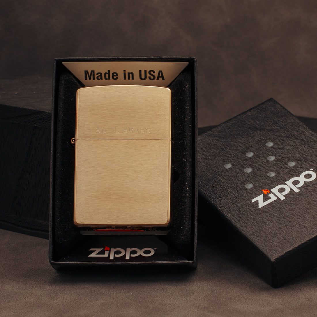 A brushed brass Zippo lighter displayed in an open black Zippo box, with the branded lid placed beside it on a dark brown textured surface.