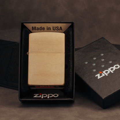 A brushed brass Zippo lighter displayed in an open black Zippo box, with the branded lid placed beside it on a dark brown textured surface.