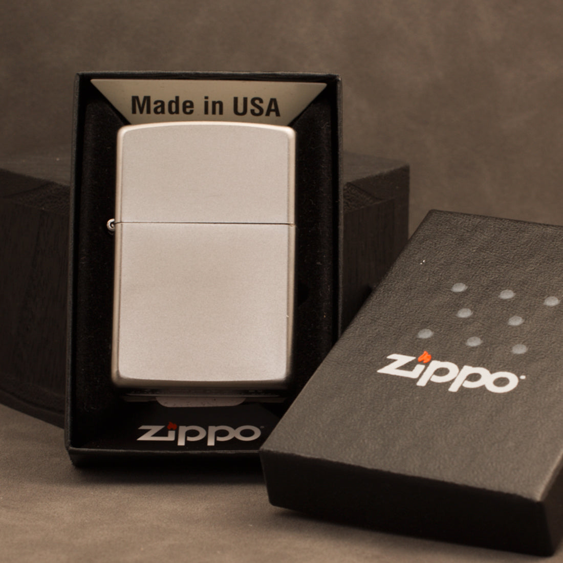A silver Zippo lighter in its original black presentation box, with the lighter lid placed beside it on a textured brown background.
