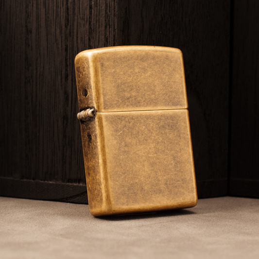 A classic rectangular lighter with a distressed, antique brass finish, standing upright on a textured gray surface against a dark wood background.