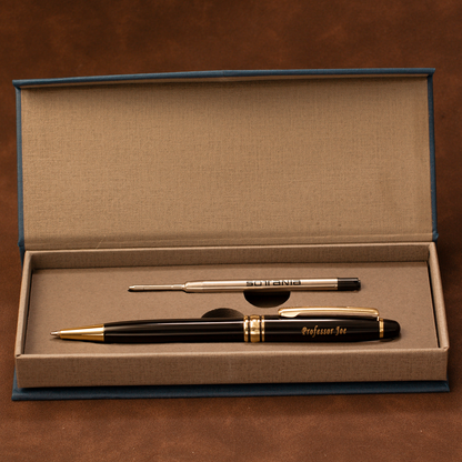 A black and gold ballpoint pen set displayed inside an open blue presentation box with a beige insert. The main pen is labeled 'Professor Joe' in gold script, and a smaller metal replacement tip is placed above it on the tray.