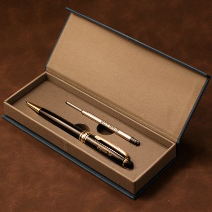 A black and gold ballpoint pen set displayed inside an open textured beige presentation box on a brown surface. The pen has gold trim and the cap is off, revealing a silver-toned metal tip or accessory.
