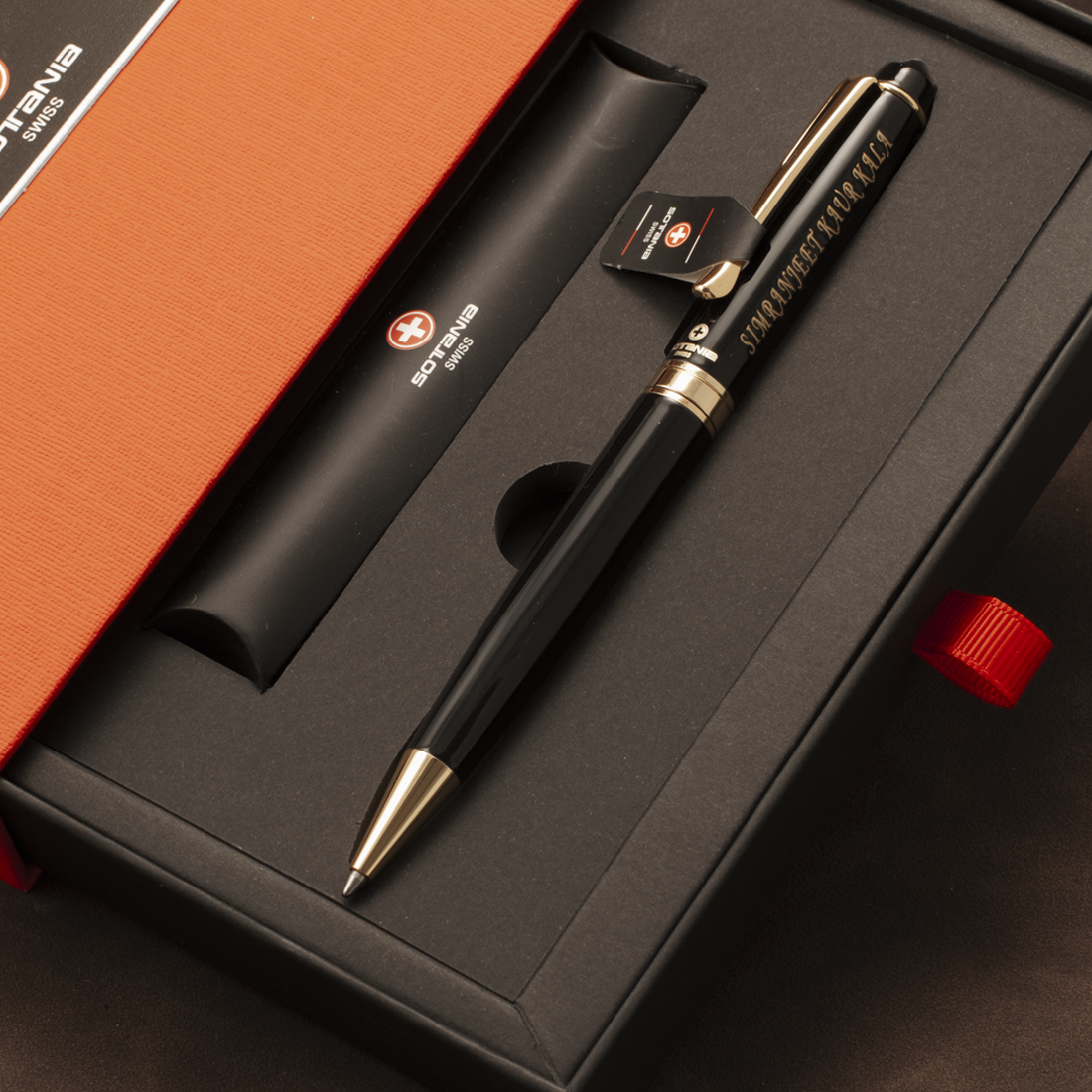 A black ballpoint pen with gold trim and Chinese characters engraved on the barrel, presented in a black gift box with an orange lid. A black cylindrical tube with a Swiss flag logo is also included.