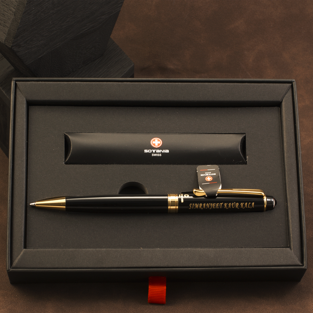 A black and gold pen presented in a black gift box, with a matching black cylindrical pouch behind it. The pen has gold trim, gold lettering reading 'SIMONSCHEEF KUVAKS', and a small Swiss cross logo with the word 'SOVENS' visible on the pouch.