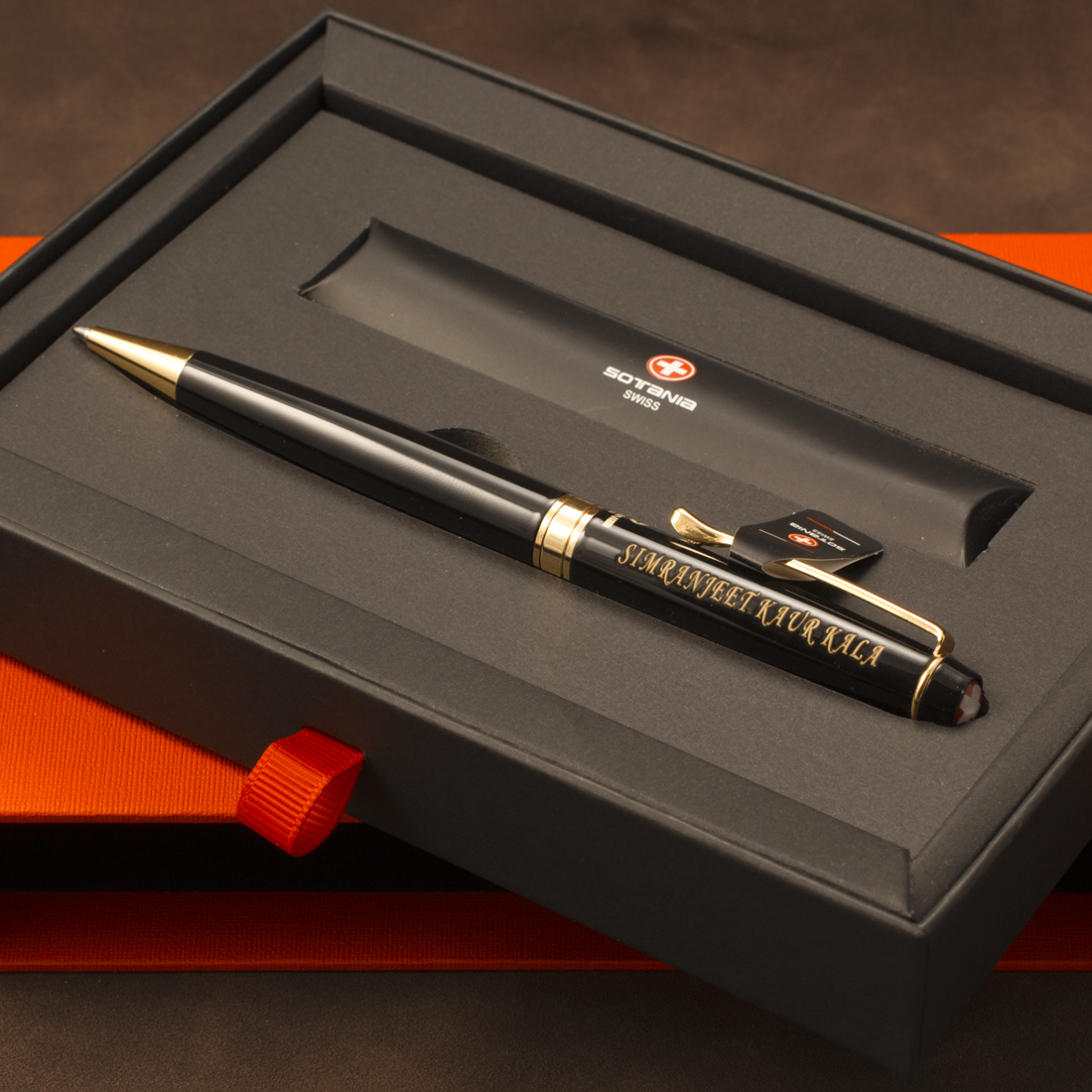 A black ballpoint pen with gold trim and gold foil lettering, presented inside a black gift box with an orange-red ribbon. The pen features a gold clip and a small black tag attached near the clip.