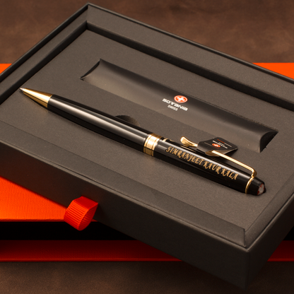 A black ballpoint pen with gold trim and gold foil lettering, presented inside a black gift box with an orange-red ribbon. The pen features a gold clip and a small black tag attached near the clip.
