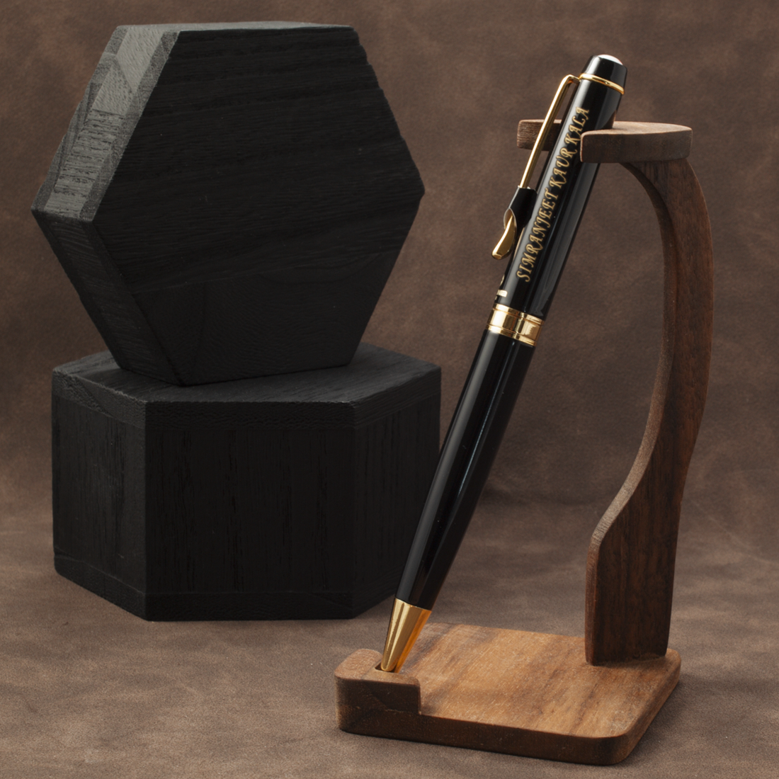 A black and gold ballpoint pen displayed on a curved wooden stand, with a dark hexagonal box behind it on a brown textured studio background.