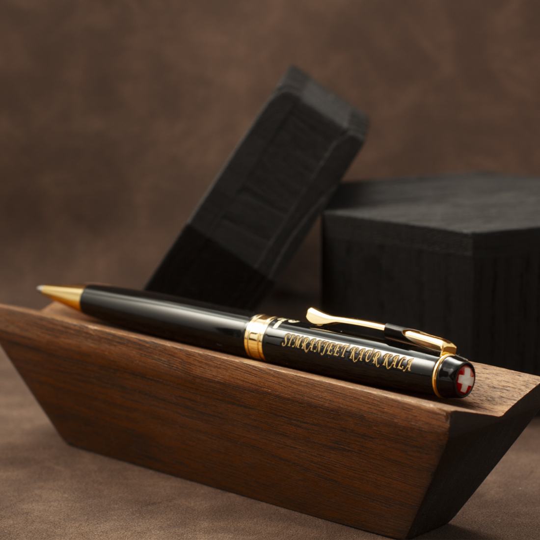 A black and gold pen displayed on a wooden stand, with two black presentation boxes behind it against a brown textured background.