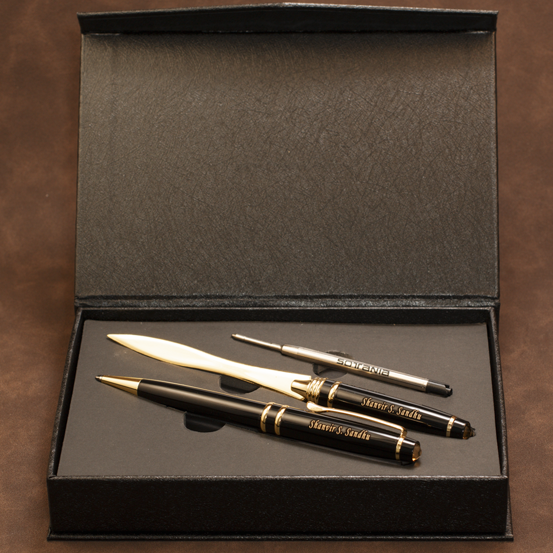 A luxury stationery set including a black and gold ballpoint pen, a silver and gold rollerball pen, and a silver and gold pencil, all displayed inside an open black presentation box on a brown textured background.