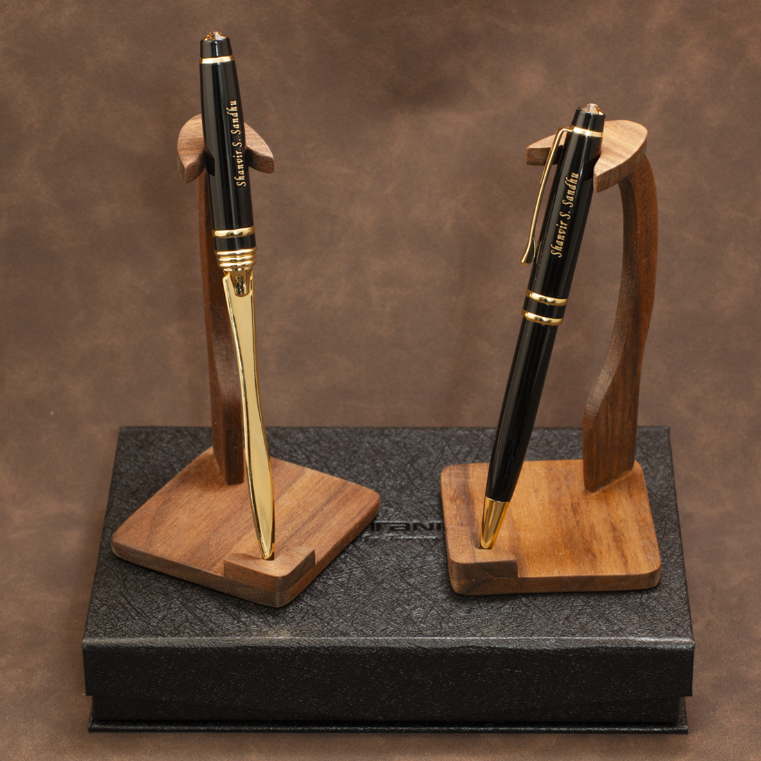 Two black and gold ballpoint pens displayed on wooden stand holders, set on top of a black presentation box against a brown textured background.