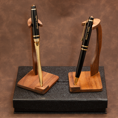 Two black and gold ballpoint pens displayed on wooden stand holders, set on top of a black presentation box against a brown textured background.