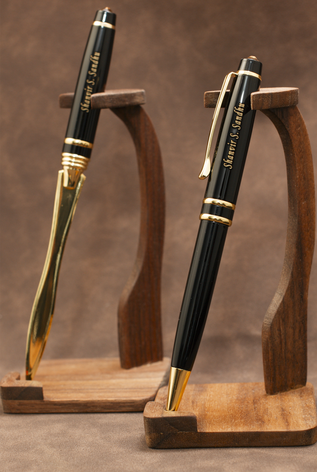 Two black and gold ballpoint pens displayed on matching wooden stands against a brown textured background. The pens are personalized with the name 'Marvin E.