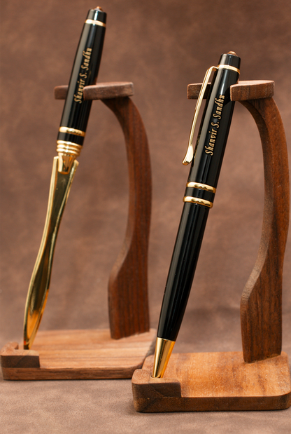 Two black and gold ballpoint pens displayed on matching wooden stands against a brown textured background. The pens are personalized with the name 'Marvin E.
