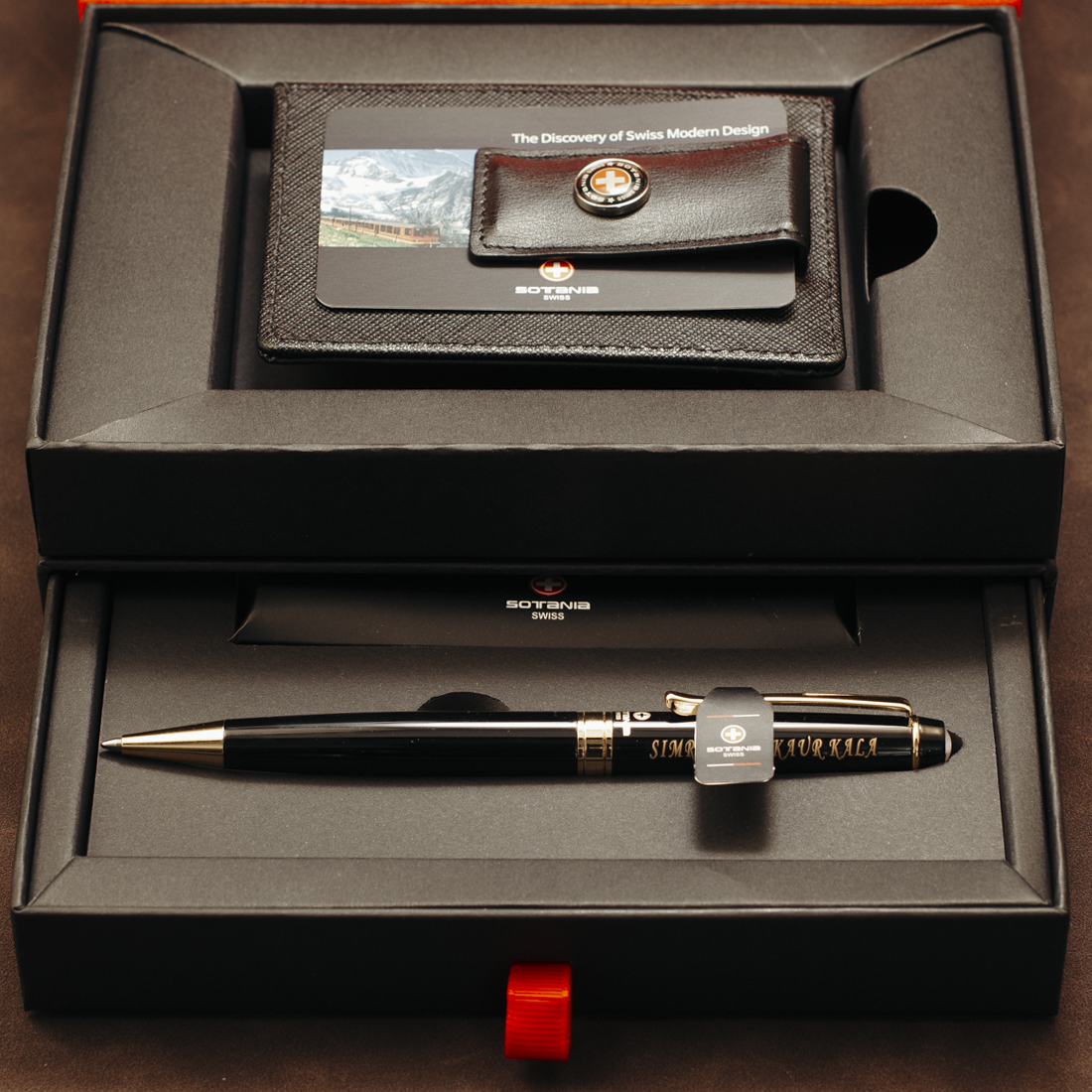 A Swiss Made pen gift set displayed in an open black presentation box. The set includes a black pen with gold trim, a black leather wallet/card holder with a mountain photo and Swiss Made logo, and a black pen sleeve.