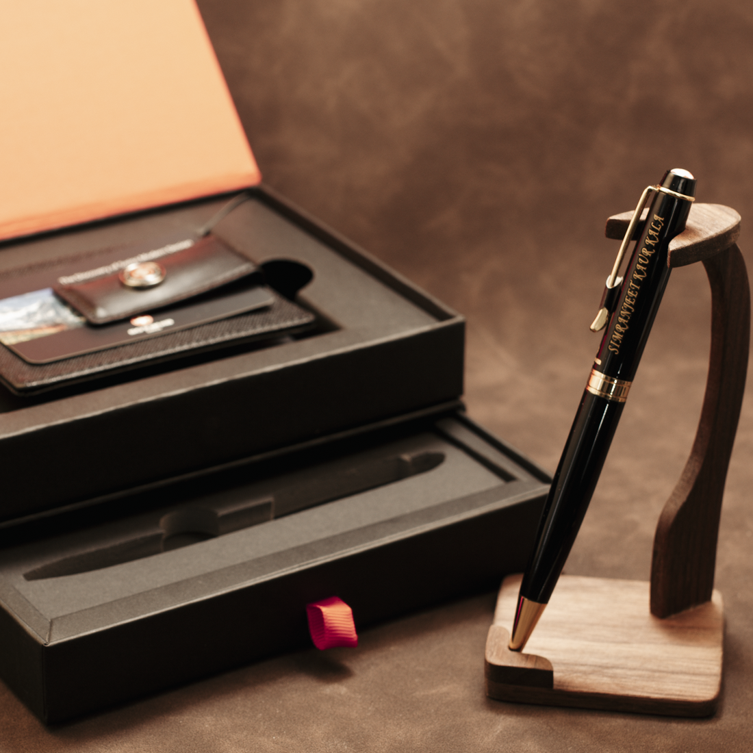 A black and gold ballpoint pen displayed on a wooden stand next to its black presentation box containing a leather wallet and cardholder, on a brown textured background.