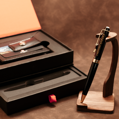 A black and gold ballpoint pen displayed on a wooden stand next to its black presentation box containing a leather wallet and cardholder, on a brown textured background.