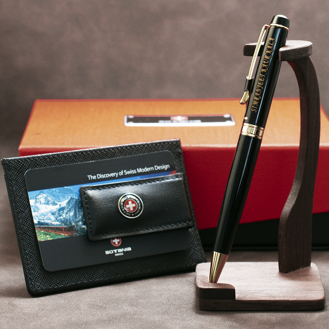 A Swiss-designed pen and card set displayed on a wooden stand with an orange and red gift box in the background. The pen is black with gold trim and the Sottus logo, resting in a wooden holder.