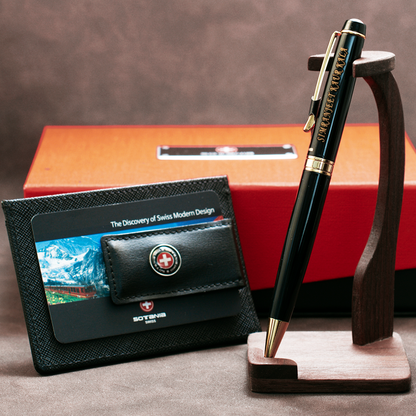 A Swiss-designed pen and card set displayed on a wooden stand with an orange and red gift box in the background. The pen is black with gold trim and the Sottus logo, resting in a wooden holder.