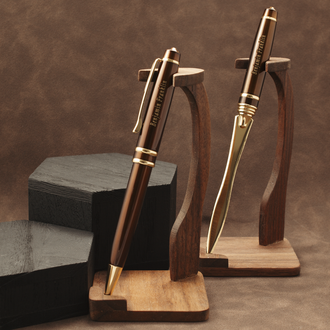 Two metallic ballpoint pens with a warm bronze-brown finish and gold accents, displayed on wooden stands against a brown textured backdrop.