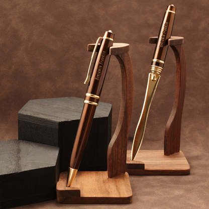 Two metallic ballpoint pens with a warm bronze-brown finish and gold accents, displayed on wooden stands against a brown textured backdrop.