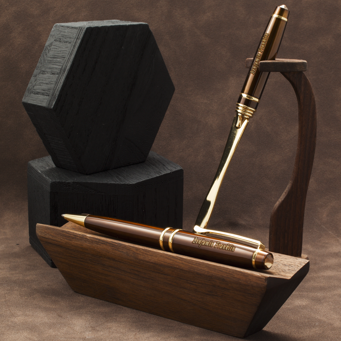 A polished brown wooden pen with gold trim and a matching wooden pen stand, displayed alongside a black hexagonal box on a brown textured background.