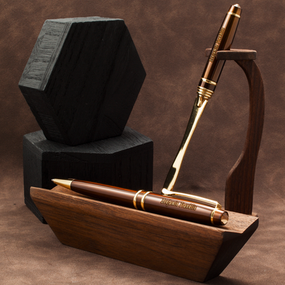 A polished brown wooden pen with gold trim and a matching wooden pen stand, displayed alongside a black hexagonal box on a brown textured background.