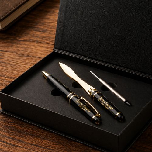 A black and gold personalized pen set displayed inside an open black presentation box on a wooden table. The set includes a black pen with gold trim and engraving, a matching gold-colored stylus, and a small silver pen tool, all resting in a black foam-lined box.