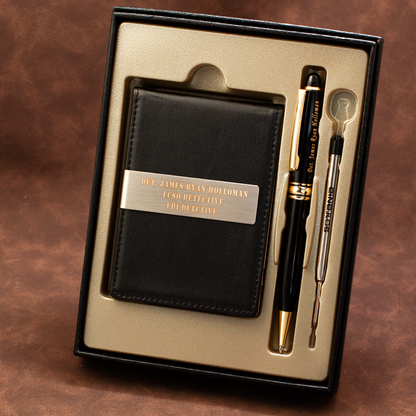 A black leather wallet set inside a black gift box with a gold interior, accompanied by a black and gold pen and a small silver tool. The wallet has a silver metal plate engraved with 'DET.