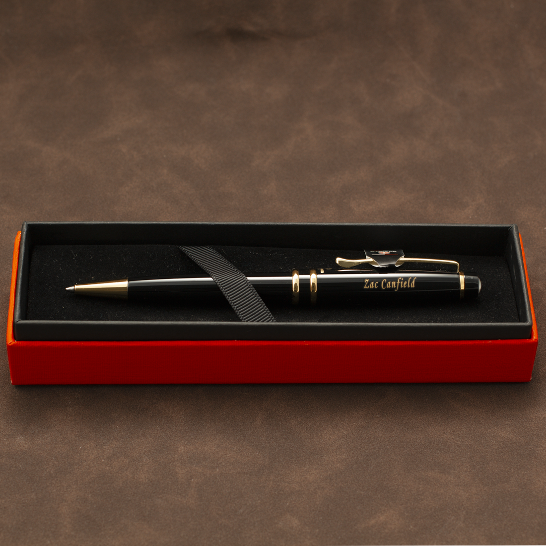 A black ballpoint pen with gold trim and clip, personalized with 'Sacramento' in gold script, presented in an open red gift box with a black interior on a brown textured background.