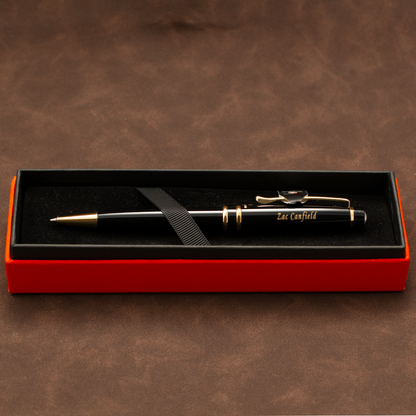 A black ballpoint pen with gold trim and clip, personalized with 'Sacramento' in gold script, presented in an open red gift box with a black interior on a brown textured background.