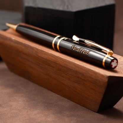 A black and gold pen with 'VisualArt' engraved on the barrel, resting on a polished wooden block.