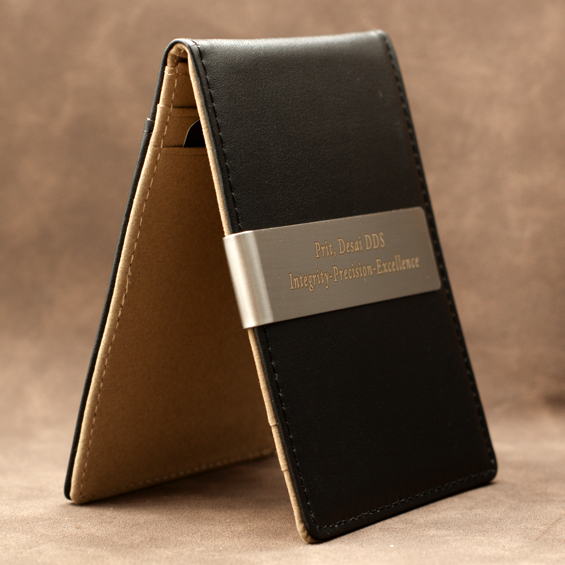 A black leather card holder wallet standing open to show its tan interior, featuring a silver metal band engraved with 'Prof. Dessi DDS Integrity-Precision-Excellence' on a brown textured background.