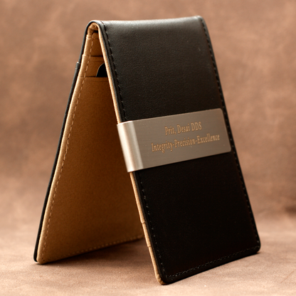 A black leather card holder wallet standing open to show its tan interior, featuring a silver metal band engraved with 'Prof. Dessi DDS Integrity-Precision-Excellence' on a brown textured background.
