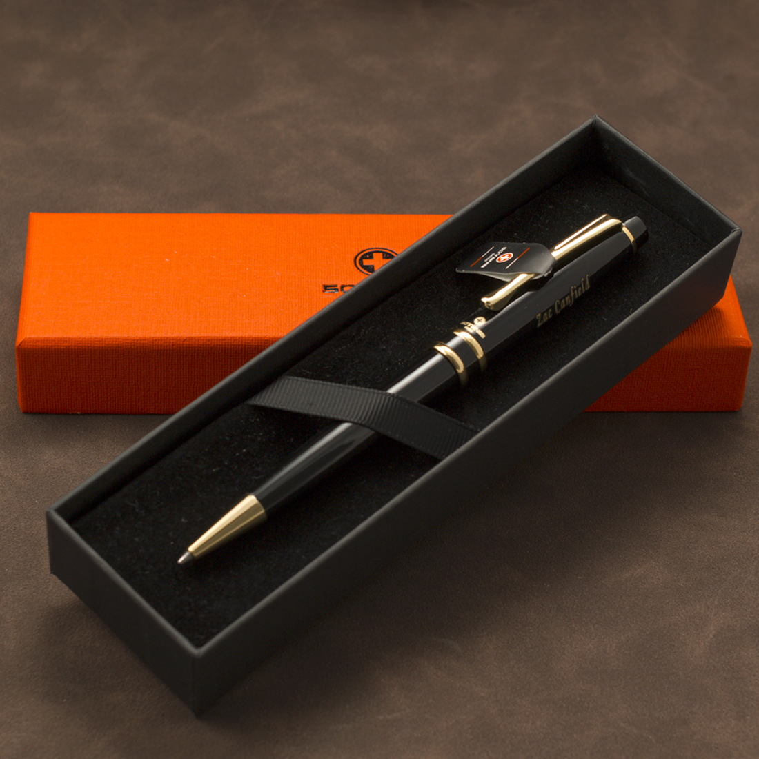 A black ballpoint pen with gold trim and a black clip, presented in an open black gift box with a black ribbon. A bright orange rectangular box sits behind it on a brown textured surface.