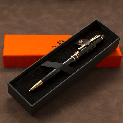 A black ballpoint pen with gold trim and a black clip, presented in an open black gift box with a black ribbon. A bright orange rectangular box sits behind it on a brown textured surface.