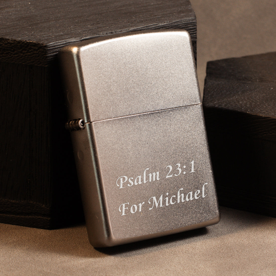 A brushed silver metal lighter with engraved white text reading 'Psalm 23:1 For Michael', resting against dark wooden boxes on a neutral brown surface.