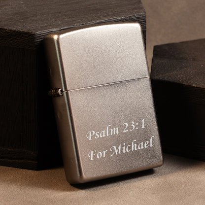 A brushed silver metal lighter with engraved white text reading 'Psalm 23:1 For Michael', resting against dark wooden boxes on a neutral brown surface.